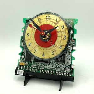 May include: A clock made from a repurposed computer hard drive circuit board. The clock face is a round, cream-coloured disc with black numbers and hands. The centre of the clock face is a red circle with a gold-coloured gear.