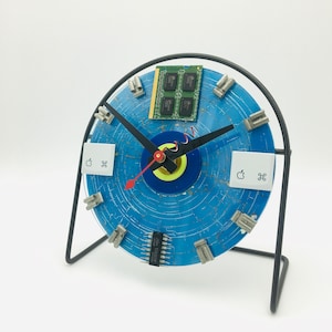 May include: A blue and silver clock made from recycled computer parts. The clock face is a blue disc with a silver ring around the edge. The clock hands are black and silver. The clock is mounted on a black metal stand.