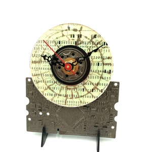 May include: A clock made from a repurposed computer hard drive. The clock face is a circular piece of clear plastic with a cracked pattern. The clock hands are black and red. The clock is mounted on a metal base that resembles a circuit board.