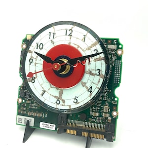 May include: A clock made from a repurposed computer hard drive. The clock face is white with black numbers and a red and blue target in the centre. The clock hands are black. The hard drive is green with gold connectors and the text "ASSEMBLY MADE IN MALAYSIA".