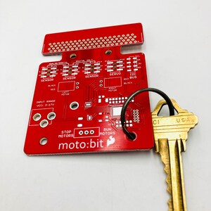 May include: A red moto:bit circuit board with various electronic components and text, including "SENSOR" and "RUN MOTORS." A key with "USA" stamped on it is attached to the board with a metal ring. The board has multiple holes and connection points.