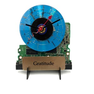 May include: A blue and black clock crafted from recycled computer components. The clock face is a repurposed CD with a black centre and blue outer ring. The clock hands are black and red. The clock is mounted on a green circuit board with a gold plaque that reads "Gratitude".