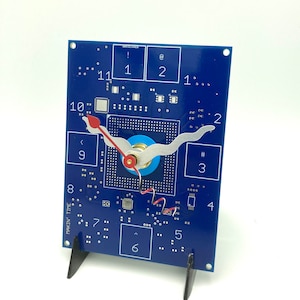 May include: A blue circuit board clock with red and silver hands. The clock face is made up of a circuit board with numbers 1 through 12 printed on it. The clock is on a black stand. The text "MAKIN' TIME" is printed on the side of the clock.