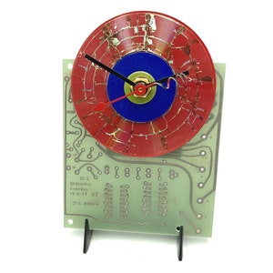 May include: A red, blue, and gold clock with a circuit board base. The clock face is a red vinyl record with a blue and gold center. The clock hands are black and red. The circuit board base has the text "BC-2 Beadwall Control 11-3-77 VT JIC AREA->".