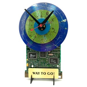 May include: A clock made from a recycled computer circuit board and a vinyl record. The clock face is blue, green, and turquoise. The clock hands are black and red. The clock is mounted on a stand with a gold plaque that reads "WAY TO GO!"