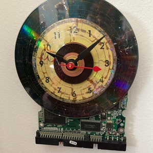 May include: A clock made from a recycled CD and a computer circuit board. The clock face is a yellowed, cracked, and worn CD with black hands and red second hand. The numbers are black and the background is a dark brown circle. The circuit board is green with black text and a black connector.