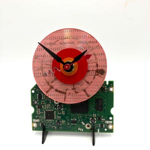 May include: A pink clock with a red centre and black hands. The clock face is made from a computer hard drive disk with the text "COBOL SOURCE PROGRAM" printed on it. The clock is mounted on a green circuit board.