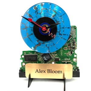 May include: A blue vinyl record clock with black hands and a red second hand. The clock is mounted on a green circuit board with the text "DC© 2005" and "SAMSUNG" visible. A gold plaque with the text "Alex Bloom" is attached to the base.