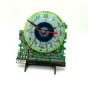May include: A clock made from a recycled computer circuit board with a green and blue face. The clock face has a cracked, distressed look and features a blue and green eye design.