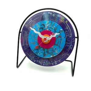 May include: A clock made from a repurposed vinyl record with a blue, purple and pink colour scheme. The clock hands are silver and red, and the clock is mounted on a black metal stand.