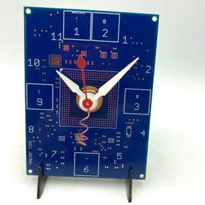 May include: A blue circuit board clock with white hands and a red second hand. The clock face is a square with numbers 1 through 12 around the edge. The clock is on a black stand. The text "MAKIN' TIME" is printed on the side of the clock.