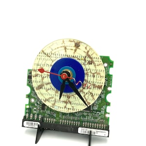 May include: A clock made from a computer hard drive with a blue and turquoise face and black hands. The clock is mounted on a green circuit board with black stand.