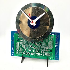 May include: A clock made from a recycled CD with a green circuit board base. The clock face is black and gold with a white and red hand.