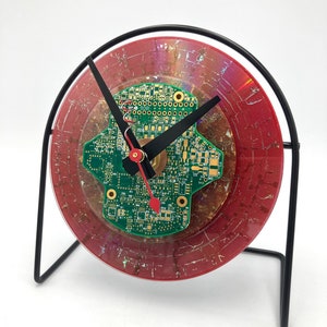 May include: A red and green clock with a circuit board face and a black metal stand.