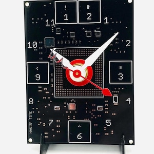 May include: A black circuit board clock with white numbers and red and white hands. The clock is on a black stand and has the text "MAKIN' TIME" on the side.