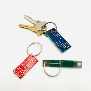 May include: Three keychains made from circuit boards. One is blue with the text "0.00 0000", one is red with the text "SCL CLK", and one is green with no text. All three keychains have silver key rings.