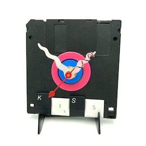 May include: A clock made from a black floppy disk with a pink and blue target face. The clock hands are made from a white floppy disk and a red arrow. The clock is sitting on a stand made from three black keys labeled "K", "I", and "S".
