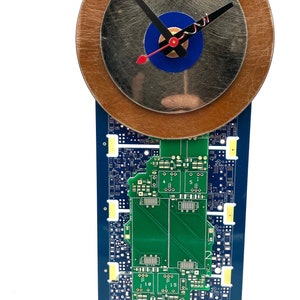 May include: A clock made from recycled circuit boards and metal. The clock face is a silver disc with a blue center and black hands. The clock is mounted on a stand made from circuit boards.