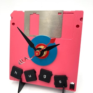 May include: A pink floppy disk clock with a blue and red clock face. The clock hands are black and the clock is mounted on a black stand. The clock is decorated with four black keys that spell out "hugs".