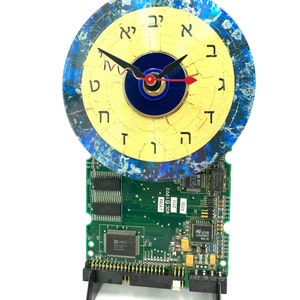 May include: A clock with a blue and yellow face, featuring Hebrew numerals, is mounted on a green circuit board. The clock hands are black and red.