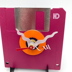 May include: A pink floppy disk with a silver metal cover and the text "HD" printed on the side. A clock face is attached to the front of the disk with orange, silver, and red hands.
