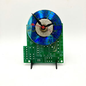 May include: A clock made from a recycled CD with a blue and green cracked surface. The clock hands are black and red. The clock is mounted on a green circuit board with black stand.