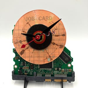 May include: A distinctive clock crafted from repurposed electronic parts. The clock face features the words "JOB CARD". Black clock hands and a red second hand are set against a circular face. The clock is mounted on a green circuit board, showcasing its unique design.