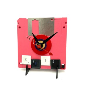 May include: A pink floppy disk clock with a red clock face and black hands. The clock is made from a repurposed floppy disk and has a stand made from black plastic. The clock face has a gold center and a purple ring around the edge. The clock is decorated with three computer keys: a white key with the letter K and the number 2, a white key with the number 5, and a black key with the letter S.