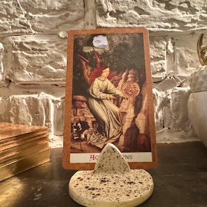 May include: A tarot card featuring an illustration of a figure with red hair and wings holding a circular object. The card is propped up by a speckled, heart-shaped ceramic piece. A stack of gold-edged cards is visible in the background.
