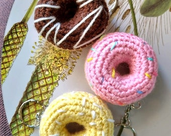 Puede incluir: Tres llaveros de donut tejidos a crochet. Uno es marrón con glaseado blanco, otro es rosa con chispas de colores y el último es amarillo con glaseado blanco.