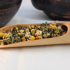 Pu&ograve; includere: Un cucchiaio di legno pieno di fiori di camomilla secchi e foglie verdi. Il cucchiaio &egrave; in legno marrone chiaro e ha una finitura naturale.