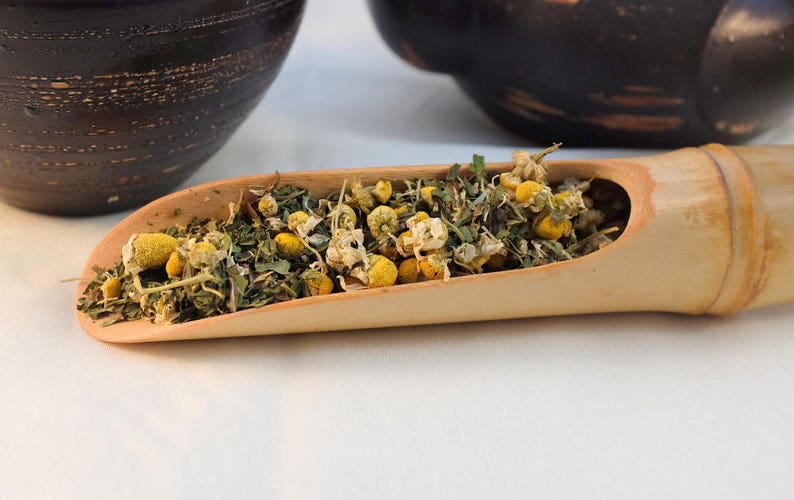 Pu&ograve; includere: Un cucchiaio di legno pieno di fiori di camomilla secchi e foglie verdi. Il cucchiaio &egrave; in legno marrone chiaro e ha una finitura naturale.