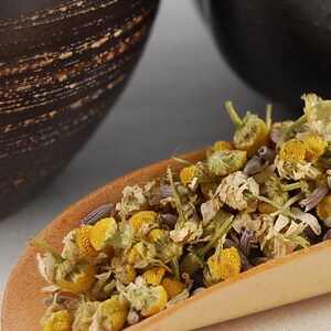 Puede incluir: Una cuchara de madera llena de flores de manzanilla y lavanda secas. Las flores son una mezcla de amarillo, blanco y morado.
