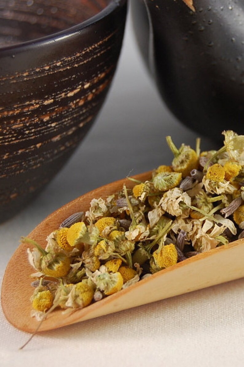Puede incluir: Una cuchara de madera llena de flores de manzanilla y lavanda secas. Las flores son una mezcla de amarillo, blanco y morado.