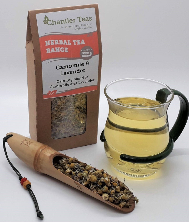 Puede incluir: Una caja de papel marr&oacute;n con una etiqueta blanca que dice "Chantler Teas Herbal Tea Range Camomile & Lavender Calming blend of Camomile and Lavender". La caja contiene flores secas de manzanilla y lavanda. Una taza de t&eacute; de vidrio est&aacute; al lado de la caja. Una cuchara de madera con flores secas de manzanilla y lavanda est&aacute; en primer plano.