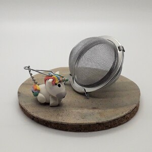 May include: A white unicorn tea infuser with a rainbow mane and tail, attached to a silver chain, sitting on a brown cork coaster next to a silver tea ball infuser.