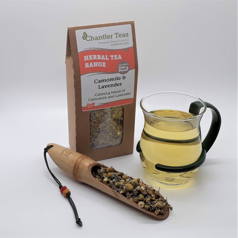 Puede incluir: Una bolsa de papel marr&oacute;n con una etiqueta blanca que dice "Chantler Teas Premium Teas Blended in Pembrokeshire HERBAL TEA RANGE Chanter's Own Blend Camomile & Lavender Calming blend of Camomile and Lavender". La bolsa est&aacute; junto a una taza de vidrio de t&eacute; y una cuchara de madera llena de flores de manzanilla y lavanda secas.