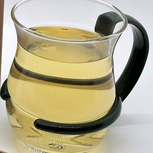 Pu&ograve; includere: Una tazza di vetro trasparente con un manico verde, riempita di un liquido giallo chiaro.