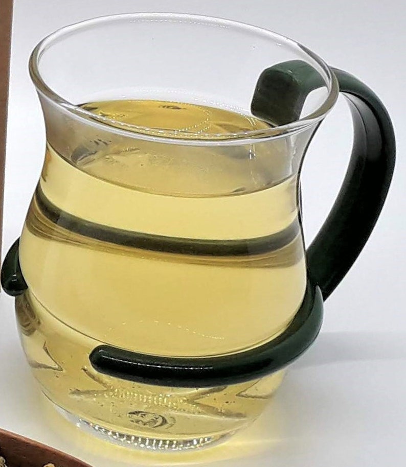 Pu&ograve; includere: Una tazza di vetro trasparente con un manico verde, riempita di un liquido giallo chiaro.