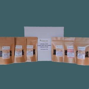 May include: A collection of six small brown paper bags containing loose leaf tea. The bags are labeled with the names of different spiced chai tea flavors, including Bangalore Rose Chai, Chill Spiced Chai, Vanilla Spiced Chai, Chandler's Spiced Chai, Chocolate Spiced Chai, and Spiced Apple Tea. The bags are arranged in a row on a white surface. The text on the bags reads "Spiced Chai Loose Leaf Tea Sample Collection by Chantier Teas."