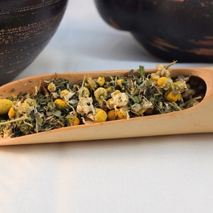 Pu&ograve; includere: Un cucchiaio di legno pieno di fiori di camomilla secchi e foglie verdi. I fiori sono bianchi con centri gialli.