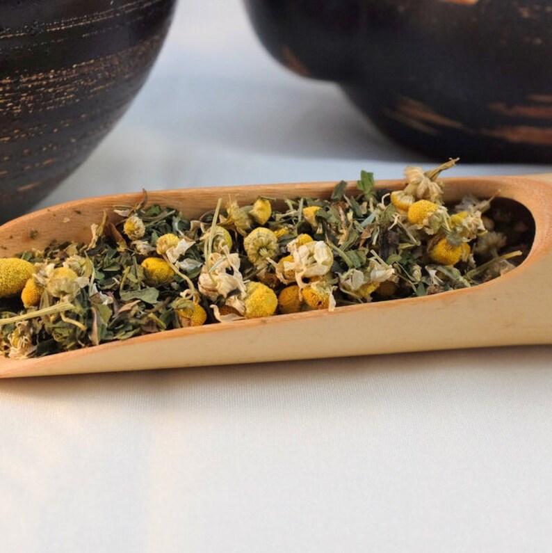 Pu&ograve; includere: Un cucchiaio di legno pieno di fiori di camomilla secchi e foglie verdi. I fiori sono bianchi con centri gialli.