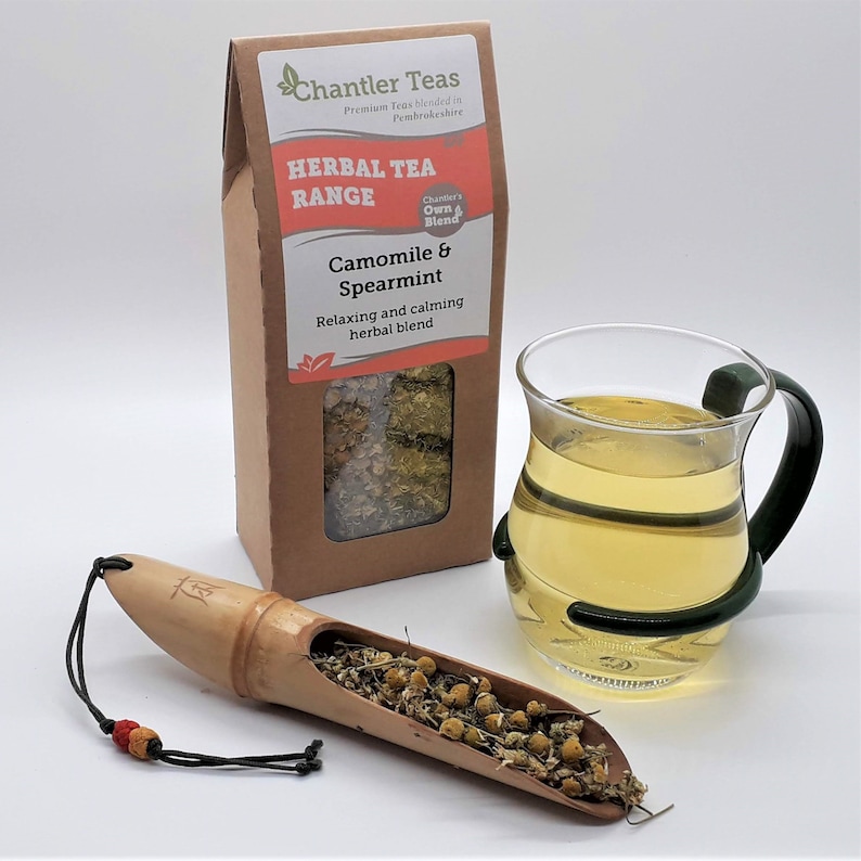Pu&ograve; includere: Una scatola di cartone marrone con una finestra che mostra t&egrave; di camomilla e menta verde essiccato. La scatola ha un'etichetta che recita "Chantler Teas Herbal Tea Range Camomile & Spearmint Relaxing and calming herbal blend". Sono inoltre visibili una tazza di t&egrave; di vetro e una paletta di legno con foglie di t&egrave;.