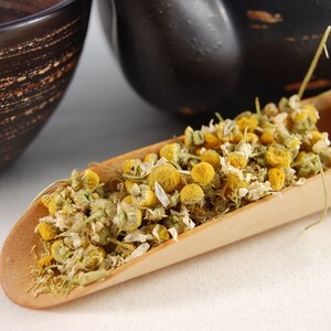 Puede incluir: Una cuchara de madera llena de flores de manzanilla secas. Las flores son de color amarillo p&aacute;lido y tienen una textura delicada y seca.