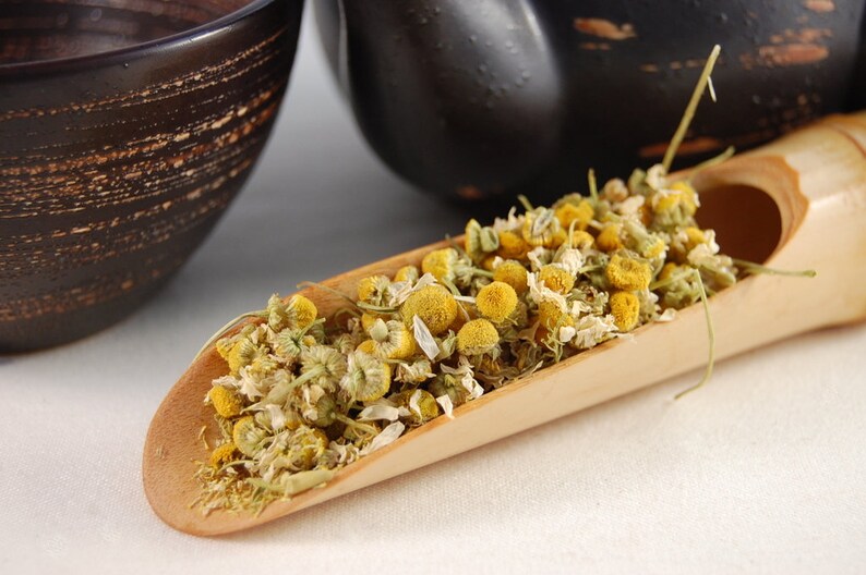 Puede incluir: Una cuchara de madera llena de flores de manzanilla secas. Las flores son de color amarillo p&aacute;lido y tienen una textura delicada y seca.