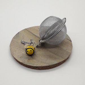 May include: A silver tea infuser shaped like a sphere with a small, yellow bee charm attached to the chain. The infuser is sitting on a beige, speckled surface.
