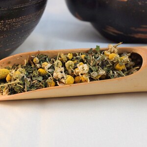 Pu&ograve; includere: Un cucchiaio di legno pieno di fiori di camomilla secchi ed erbe verdi. Il cucchiaio &egrave; in bamb&ugrave; marrone chiaro e si trova su una superficie bianca.