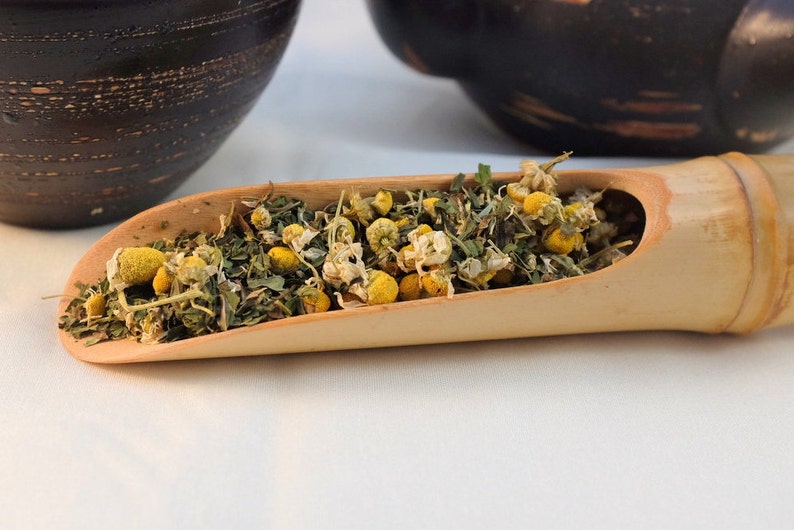 Pu&ograve; includere: Un cucchiaio di legno pieno di fiori di camomilla secchi ed erbe verdi. Il cucchiaio &egrave; in bamb&ugrave; marrone chiaro e si trova su una superficie bianca.