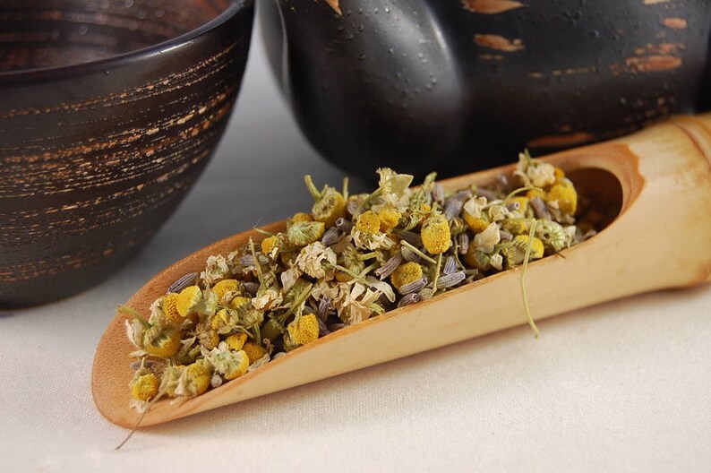 Puede incluir: Una cuchara de madera llena de flores de manzanilla y lavanda secas. Las flores son una mezcla de amarillo, blanco y morado.