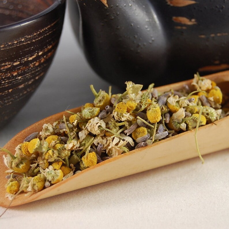 Puede incluir: Una cuchara de madera llena de flores de manzanilla y lavanda secas. Las flores son una mezcla de amarillo, blanco y morado.